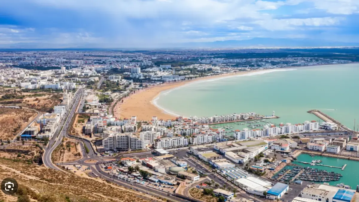 Vue aérienne d’Agadir avec baie en arc, plage dorée, marina, digues et front de mer sous ciel lumineux.