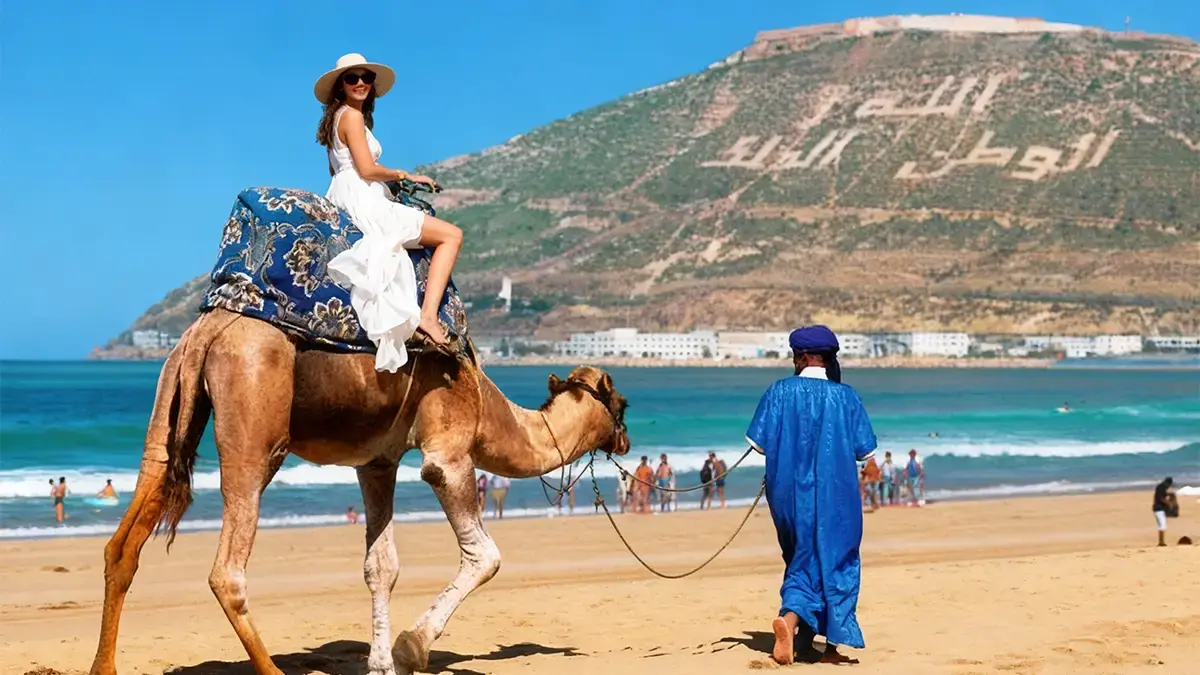 Femme en robe blanche montée sur un dromadaire guidé par un chamelier en bleu sur la plage d’Agadir, baie et colline d’Agadir Oufella en arrière-plan.