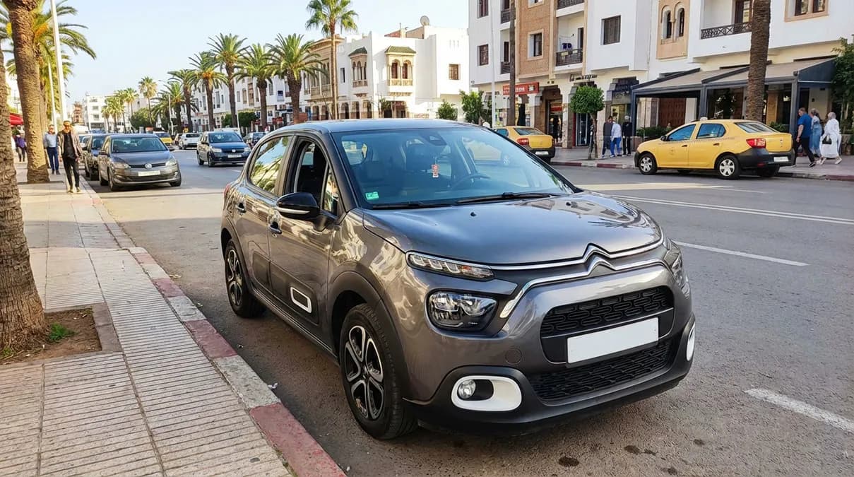 Citroën C3 Normal grise stationnée dans une rue animée d'Agadir avec palmiers en arrière-plan