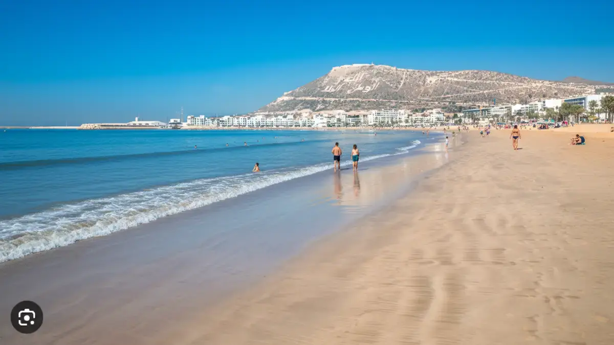Large plage d’Agadir avec sable fin, eau calme et promeneurs, panorama sur la baie et la colline inscrite surplombant la ville.